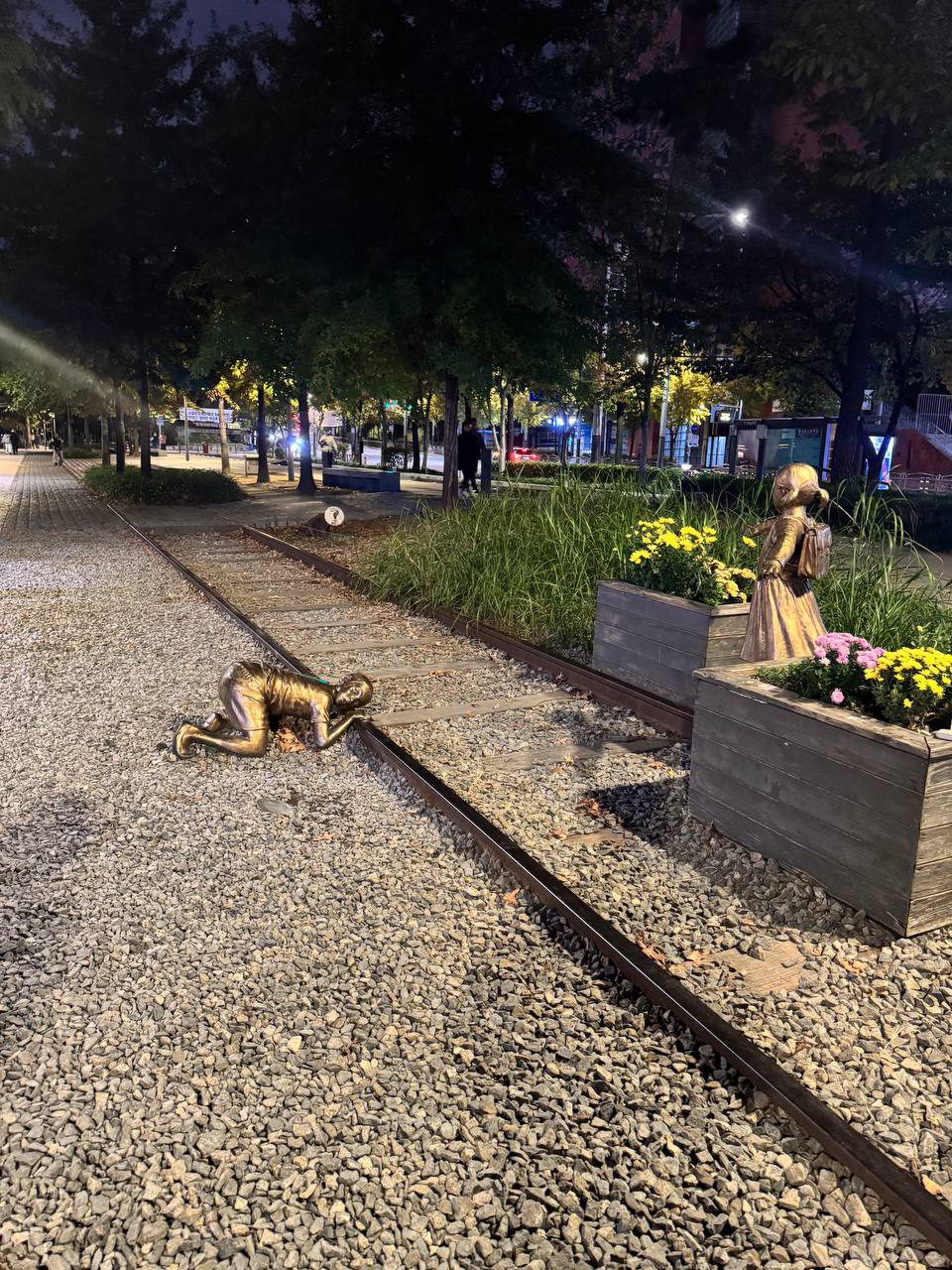 다른 방향에서 본 동상. 레일에 귀를 대고 엎드린 남자아이와, 가방을 메고 머리를 묶은 여자아이가 노란 국화와 분홍 국화가 담긴 나무 화분 옆 레일 위에 서 있다. 철로 사이로 낡은 침목이 깔려 있다