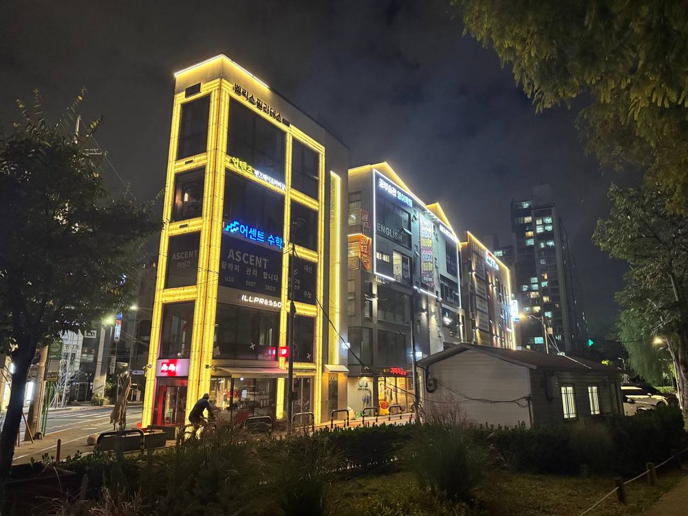 숲길의 수풀 너머로 노란 조명을 두른 건물과 옆 건물들이 이어지고, 외벽에는 수학·영어 학원 간판이 보인다