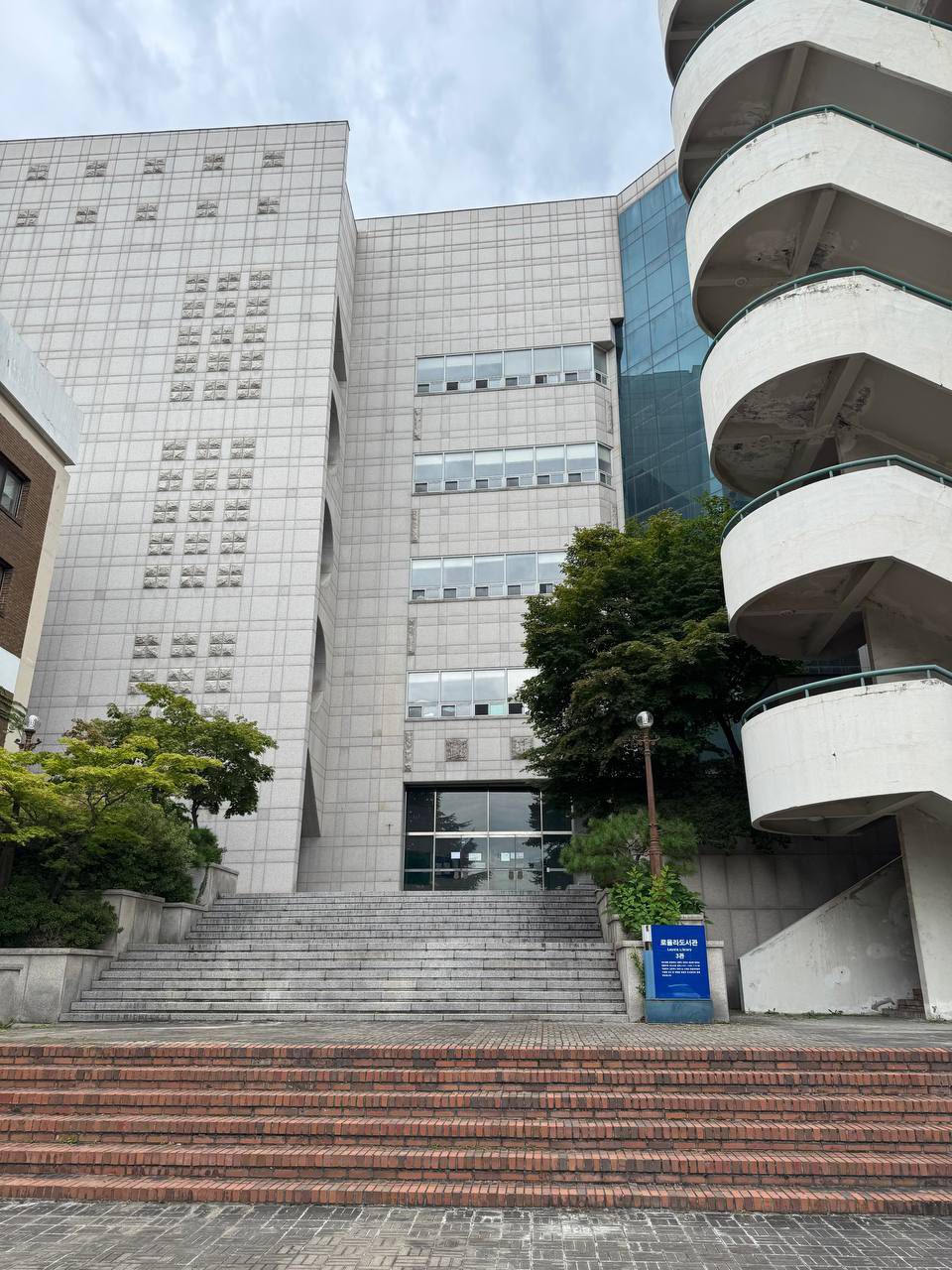 도서관 신관의 유리 출입문으로 이어지는 넓은 돌계단, 오른쪽에는 둥글게 돌아 올라가는 외부 계단과 파란 도서관 안내판이 보인다