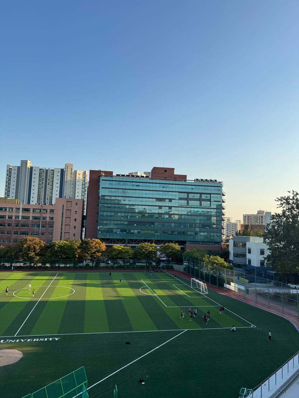 유리창이 넓게 난 건물 앞에 단풍 든 나무들이 이어지고 운동장 오른쪽에 사람들이 모여 있다