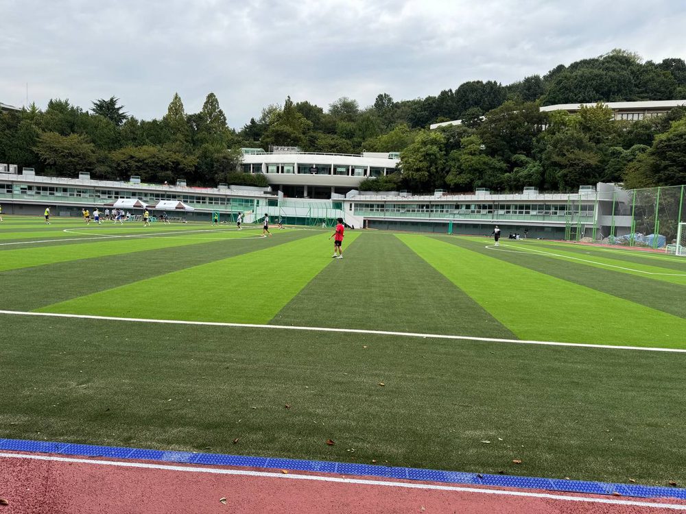 줄무늬 인조잔디와 흰 경기장 선이 펼쳐진 대운동장, 그 너머로 보이는 엠마오관