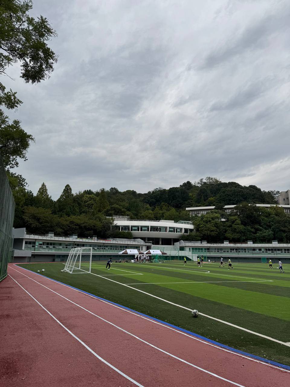 골대와 축구하는 사람들이 보이는 운동장 옆으로 붉은 트랙이 휘어지고, 건너편에는 천막과 낮은 건물이 이어진다