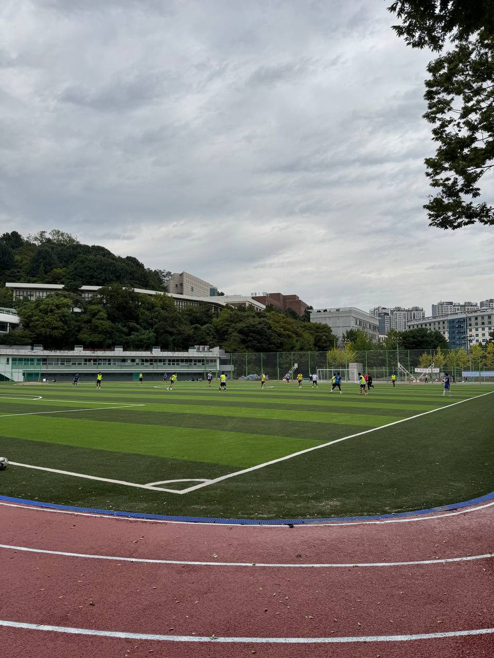 운동장 모서리의 흰 선과 붉은 트랙 너머로 축구하는 사람들, 교내 건물과 멀리 아파트가 보인다