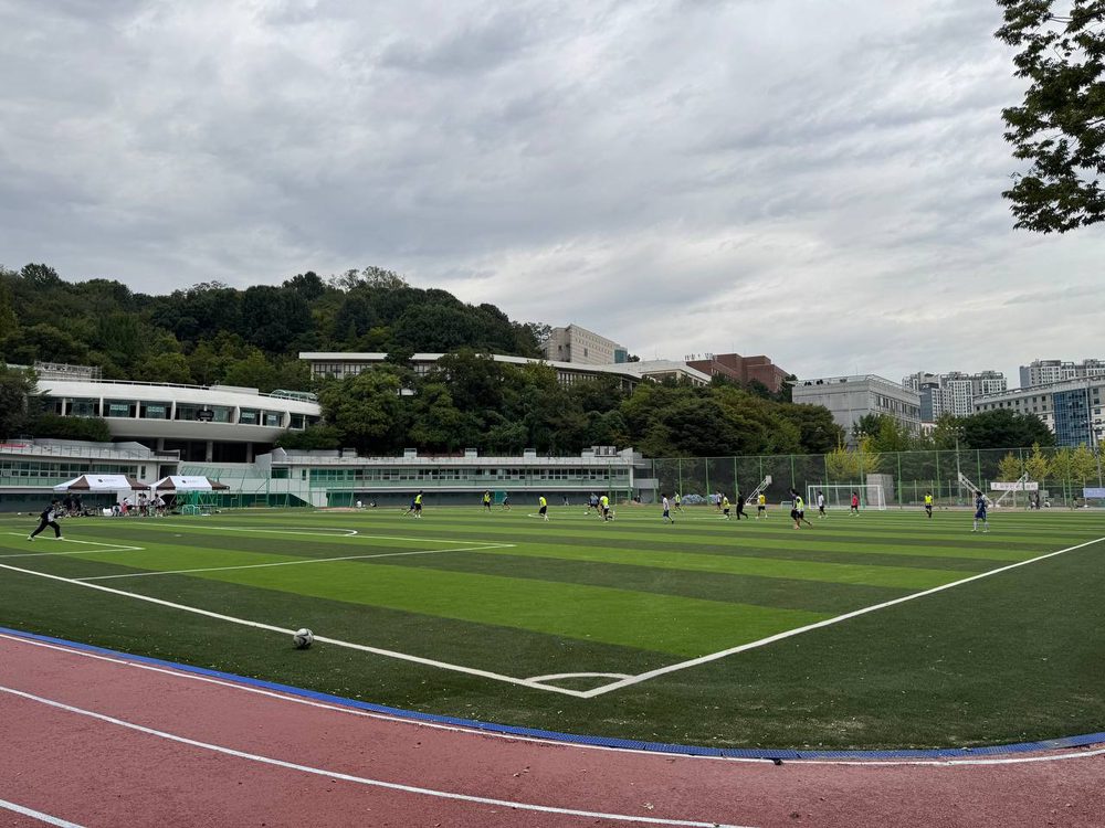 붉은 트랙 안쪽 경계선 부근에 축구공이 놓여 있고, 넓은 구장과 엠마오관, 주변 나무들이 한눈에 들어온다