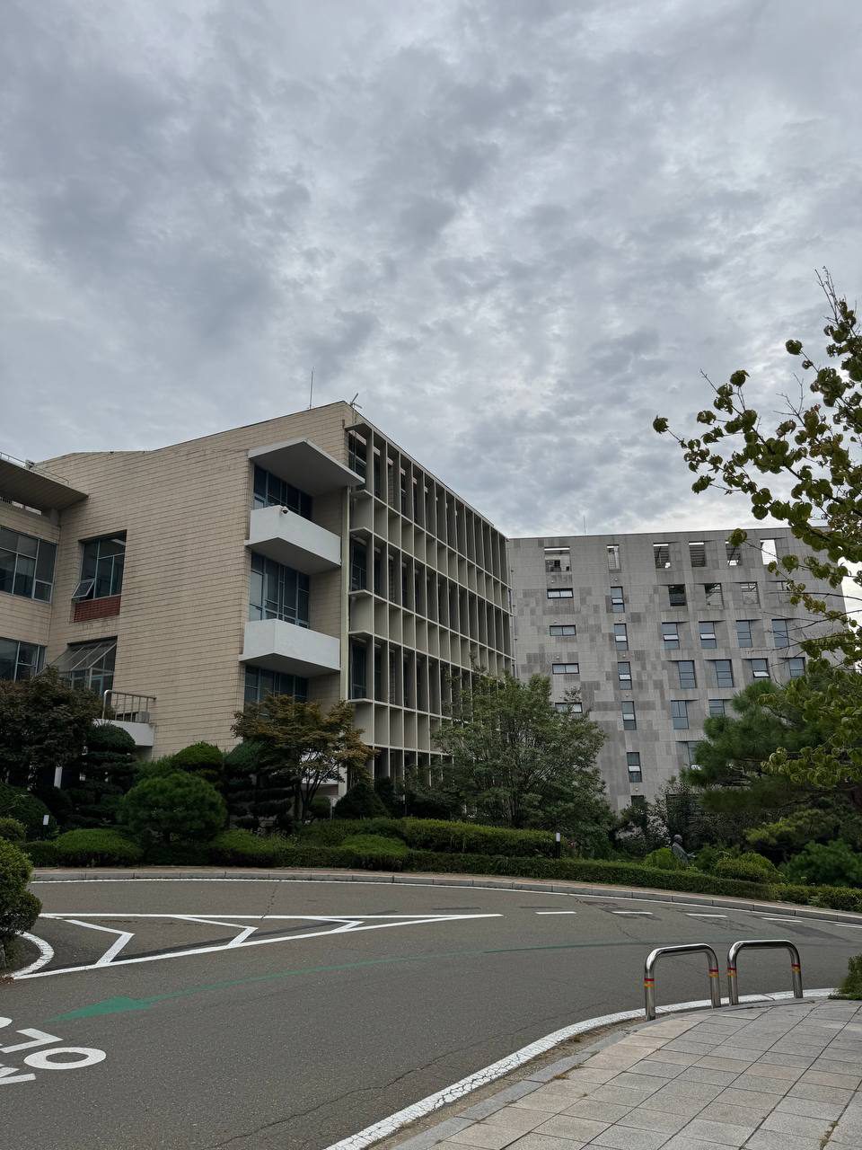 굽은 도로 건너편에 본관의 밝은 외벽과 격자 구조가 보이고 오른쪽에는 회색 건물이 서 있다