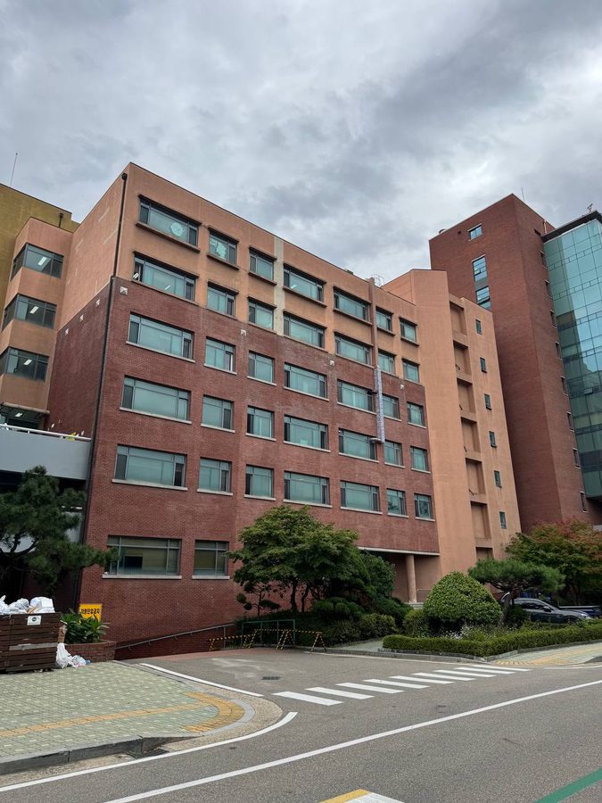 붉은 벽돌과 살구색 외벽의 캠퍼스 건물 앞에 나무와 화단이 있고 도로에는 횡단보도가 놓여 있다