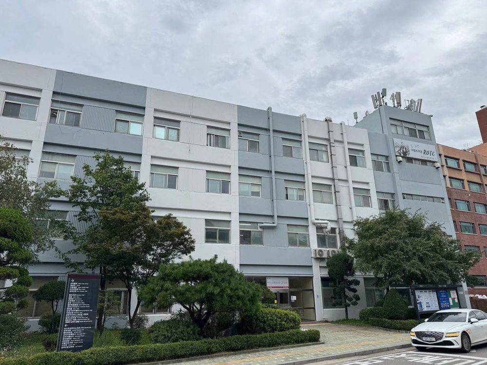 회색과 흰색 외벽의 건물 앞에 나무와 안내판이 서 있고, 건물 오른쪽 위에는 ROTC 표지가 보인다